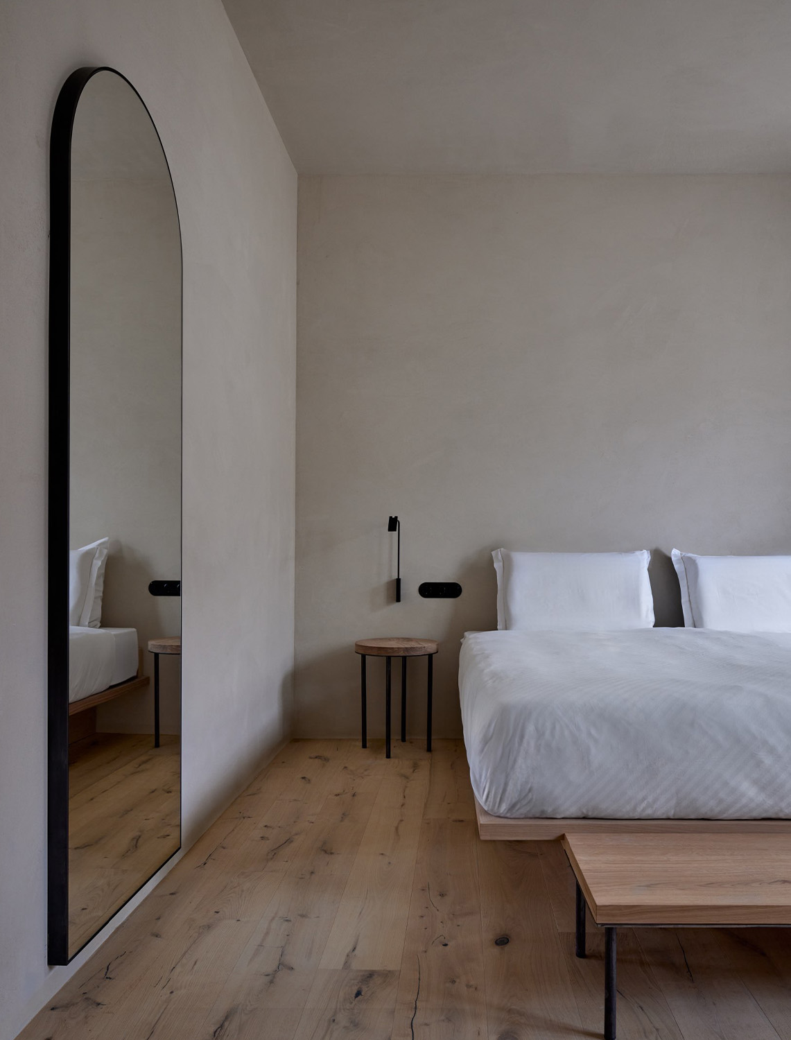 Bed with white bed linen, wooden floor, bedside table and an arch-shaped mirror on the left wall